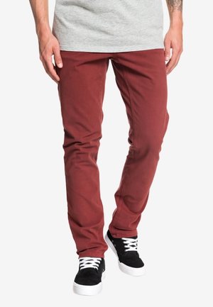 Homme portant un pantalon slim rouge rouille, des baskets noires avec des lacets blancs et une chemise gris clair, debout, les mains détendues le long du corps.