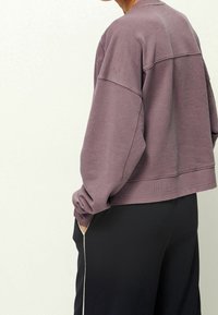 Fialová mikina s volným střihem, žebrovaným lemem a manžetami, strukturovaná tkanina, nošená s černými kalhotami s bílým bočním pruhem.