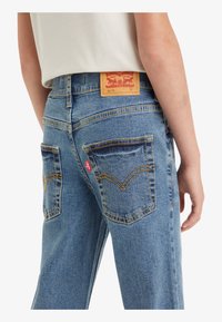 Jeans de mezclilla azul con un corte relajado, que presentan una etiqueta de cuero marrón en la cintura, costuras amarillas y dos bolsillos traseros con un diseño en forma de V.