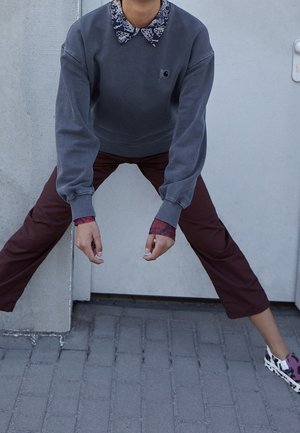 Osoba stojąca z rozstawionymi szeroko nogami i pochylona do przodu, ubrana w szarą bluzę, koszulę z wzorzystym kołnierzem, bordowe spodnie i fioletowe trampki.