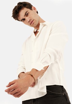 Weißes Langarmhemd mit Knopfleiste und aufgerollten Manschetten, kombiniert mit dunklen Jeans und einem goldenen Kettenarmband am Handgelenk.