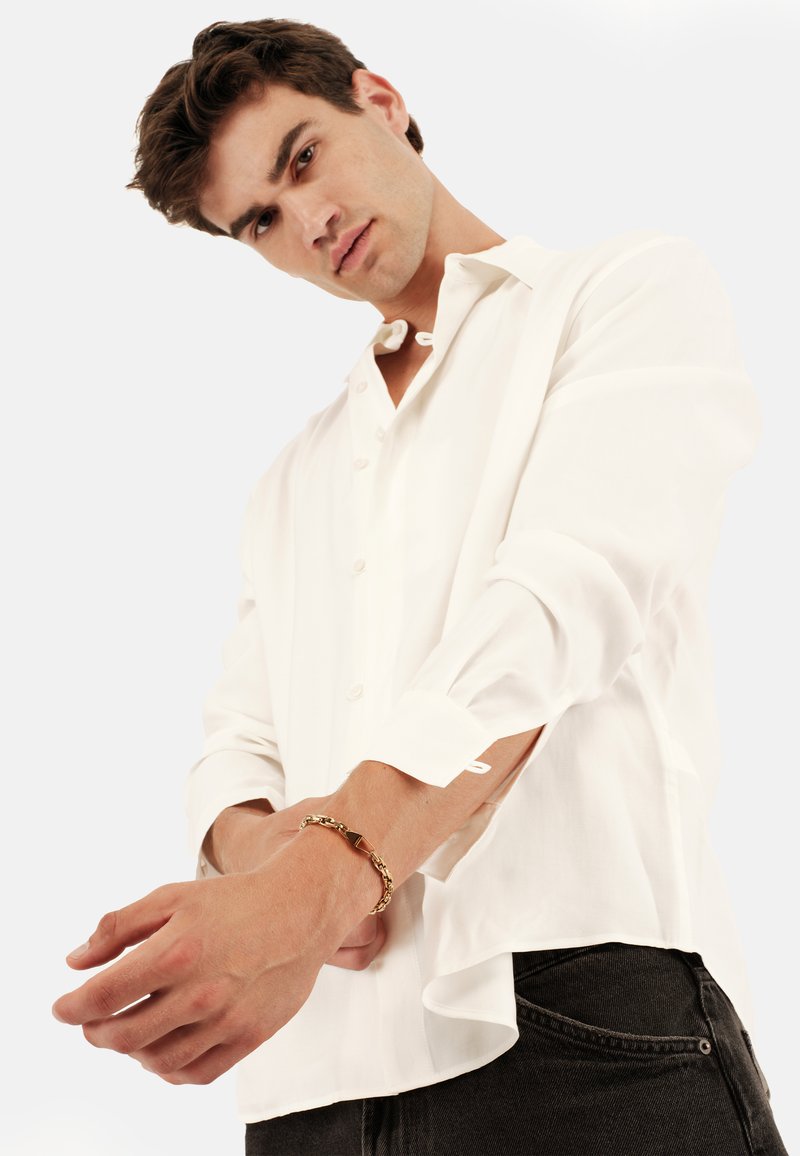 Weißes Langarmhemd mit Knopfleiste und aufgerollten Manschetten, kombiniert mit dunklen Jeans und einem goldenen Kettenarmband am Handgelenk.