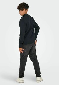 Chemise noire à manches longues côtelée associée à un pantalon en denim gris foncé. La tenue présente une coupe décontractée, des poches visibles et des baskets blanches.