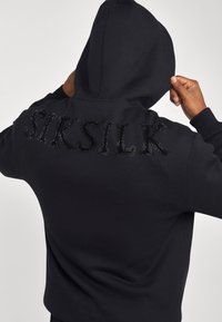 Hoodie noir avec une capuche et des lettres texturées "SIKSILK" ornées d'accents scintillants au dos. Tissu doux avec un design ajusté.
