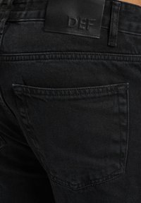 Black denim jeans with a textured finish, featuring a centered back pocket and a black leather waistband label embossed with "DEF."