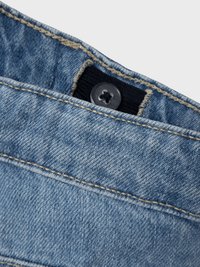 Svijetloplave traperice s tamnim gumbom i detaljem od crne tkanine. Imaju kontrastne šavove i prikaz bliže trake za struk.