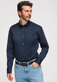 Homme portant une chemise bleu marine à boutons rentrée dans un jean bleu clair avec une ceinture marron foncé, regardant sur le côté.