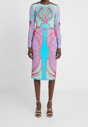 Kobieta w sukience za kolano z długim rękawem, ozdobionej misternym abstrakcyjnym wzorem w pastelowym niebieskim, różowym i czerwonym kolorze, w połączeniu z białymi sandałami na obcasie z paskami.