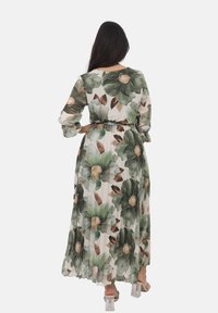 Robe maxi à fleurs avec un design plissé. Elle présente de grandes feuilles vertes et de doux accents bruns sur un fond crème. Manches longues et taille ceinturée.