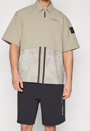 Man draagt een lichtbeige korte mouwen shirt met ritssluiting en Terrex-logo, en een zwarte korte broek met Terrex-bedrukking op het been.