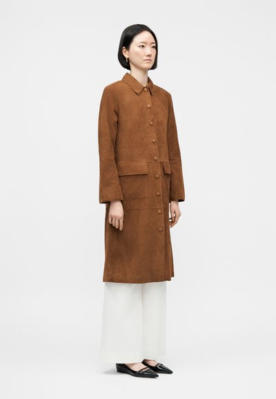 Γυναίκα που στέκεται με ένα μακρύ σουέντ παλτό σε καφέ χρώμα, κουμπωμένο, πάνω από λευκό παντελόνι φαρδύ στα πόδια και μαύρα μυτερά παπούτσια, μπροστά από απλό φόντο.