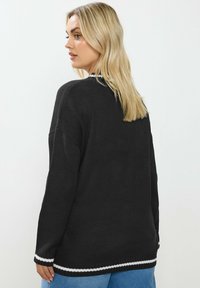 Musta neulepaita rennolla istuvuudella, jossa on valkoisia raidallisia yksityiskohtia hihansuissa ja helmassa, teksturoitu kangas ja pudotetut olkaimet.