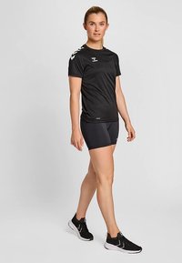 Camiseta deportiva negra de manga corta, con logo blanco y detalles en forma de chevrón en los hombros. Combinada con pantalones cortos negros y zapatillas para correr.