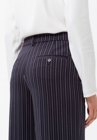 Pantalons à rayures marines avec une poche arrière boutonnée, coupe ajustée et texture lisse. Associés à une chemise blanche à manches longues ajustée.
