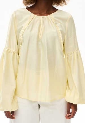 Osoba ubrana w bladoróżową bluzkę z długimi, bufiastymi rękawami i dekoltem w kształcie dziurki na kluczyk z guzikami, połączoną z jasnymi spodniami.