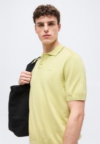 Νεαρός άνδρας που φορά ανοιχτοπράσινο πόλο με κοντά μανίκια, κρατώντας μια μαύρη τσάντα ώμου, κοιτάει μπροστά σε λευκό φόντο.