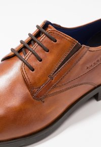 Chaussure de ville en cuir marron avec une texture lisse, bout rond, couture décorative et lacets marron. Dispose d'une semelle en caoutchouc noire.
