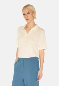 Cream-colored, short-sleeve polo shirt with a relaxed fit and a v-neck. Paired with light blue, tailored trousers. Smooth texture.