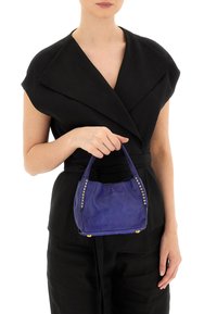 Piccola borsa in pelle viola di forma arrotondata, con dettagli borchiati lungo i lati e doppio manico, tenuta da una persona in un outfit nero.