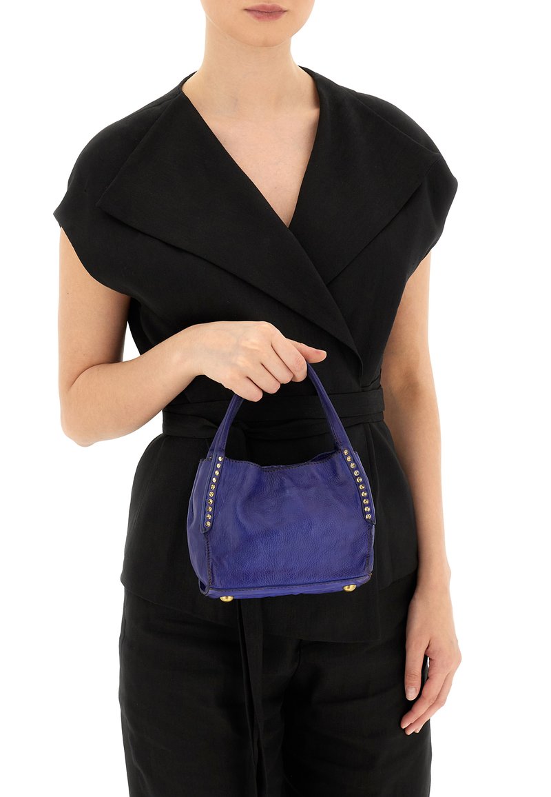 Piccola borsa in pelle viola di forma arrotondata, con dettagli borchiati lungo i lati e doppio manico, tenuta da una persona in un outfit nero.