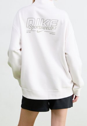 Personne portant un sweat-shirt blanc Nike Sportswear et un short noir, dos face à la caméra, avec un texte et un logo au dos du sweat-shirt.