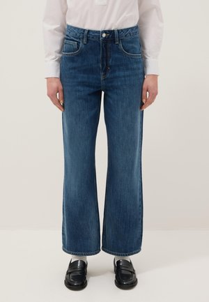 Persoon die mid-rise rechtvallende blauwe spijkerbroek, wit shirt en zwarte loafers draagt, staand tegen een effen lichte achtergrond.