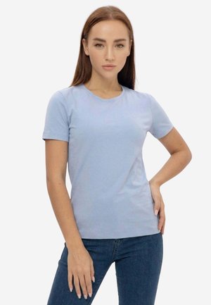 Giovane donna con capelli castani lisci che indossa una maglietta azzurro chiaro a maniche corte e jeans blu scuro, posa con una mano sui fianchi.