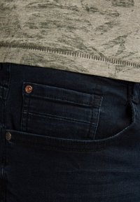 Tummansiniset farkut näyttävät selkeiltä tikkauksilta, näkyvästä taskusta ja kuparinvärisestä niitistä. Kankaan pinta on sileä.