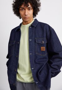 Denim jacket in dark blue with metal buttons, two front pockets, and a brown leather logo patch. Worn over a light green T-shirt.