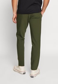 Schmal geschnittene olivgrüne Hose mit Seitentaschen, aus einem glatten Stoff, kombiniert mit off-white Sneakern mit roten Akzenten.