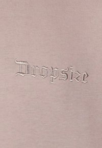 Lys pink stof med en glat tekstur, der har broderet teksten "Dropsize" i en stiliseret, middelalderlig skrifttype.