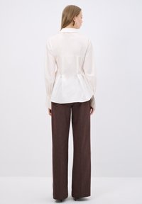 Wit shirt met een geplooid achterpand, lange mouwen en een kraag, gecombineerd met hoge, wijde bruine broeken in een gestructureerde stof.