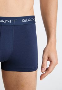 Nahaufnahme einer Person, die marineblaue Boxershorts mit gebrandetem Bund und sichtbarem Arm vor einem einfarbigen Hintergrund trägt.