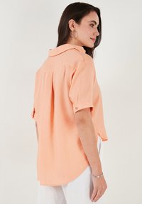 Short-sleeve, light orange blouse with a pointed collar, buttoned cuffs, and a curved hem. Soft texture, relaxed fit.