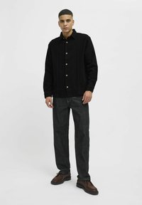 Camisa chaqueta de pana negra con cierres de botón, combinada con jeans oscuros y zapatos marrones con cordones. Fondo liso, modelo masculino de pie.