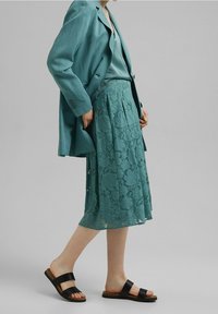 Blazer azul verdoso y falda de encaje a juego con patrón floral, combinados con sandalias negras. Tejido suave, falda hasta la rodilla con pliegues.