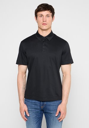 Ung mand iført en ensfarvet sort kortærmet polo og blå jeans, står foran en lys grå baggrund.