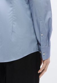 Hele sinine kraega särk pikkade varrukatega, millel on nööpidega varrukaotsad ja sile tekstuur. Alaääre lõige on kumer. All paistab must riietus.