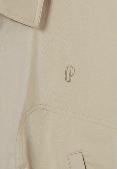 Tessuto beige con una superficie liscia, presenta un colletto e un delicato logo ricamato "CP". Include cuciture discrete e una tasca a patta.