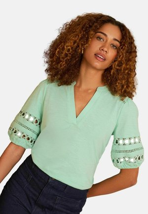 Göndör hajú nő, aki mentazöld blúzt visel díszes kivágásokkal az ujjain és sötét magas derekú farmert, fehér háttér előtt pózol.