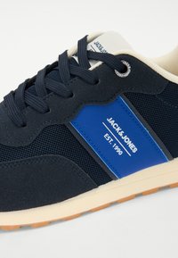 Tengeri és kék sportcipők hálós és velúr felsőrésszel, fehér logóval, kerek fűzőkkel és krém színű, texturált gumitalppal.