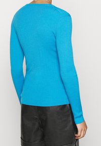 Personne portant un pull côtelé à manches longues bleu vif et un short en cuir noir, vue de dos sur un fond uni.