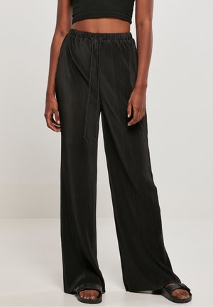 Vrouw die zwarte, wijde, geplooide broek met trekkoord in de taille, een zwart cropped topje en zwarte sandalen met open teen draagt, staand tegen een effen achtergrond.