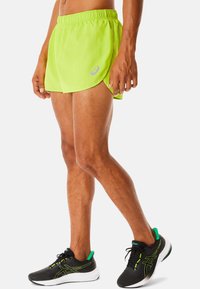 Pantalones cortos de running verde neón con una textura ligera y suave, y una cintura elástica. Combinados con zapatillas deportivas negras y verdes.
