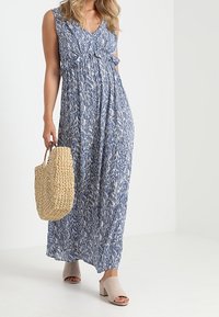 Maxi dress biru dan putih dengan pola, leher v, dan detail tali di pinggang. Dipadukan dengan tas jerami dan sandal hak blok berwarna terang.