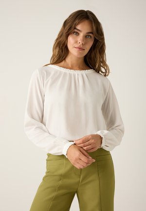 Weiße Bluse mit gerafftem Ausschnitt und elastischen Bündchen, kombiniert mit grünen Hosen mit Gesäßtaschen und taillierter Passform.