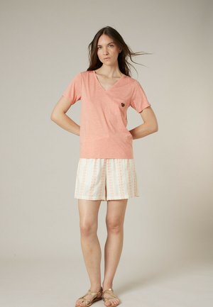 Femme aux longs cheveux bruns portant un T-shirt rose à col en V, un short blanc à motifs et des sandales beiges, debout devant un fond uni.