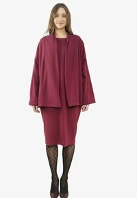 Kobieta ubrana w luźną bordową sukienkę i kardigan, z czarnymi rajstopami w wzory oraz brązowymi szpilkami, stoi na białym tle.