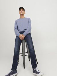 Vaaleansininen collegepaita, jossa on ribbauskät, tummansiniset slim-fit farkut ja laivastonsiniset lenkkarit, joissa on valkoiset nauhat, istuu mustalla metallijakkaralla.