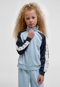 Noor tüdruk lokkis juustega, kes kannab helesinist ja tumesinist dressipluusi ševroontriipudega, hoides lukuklambrit oma kaela juures.